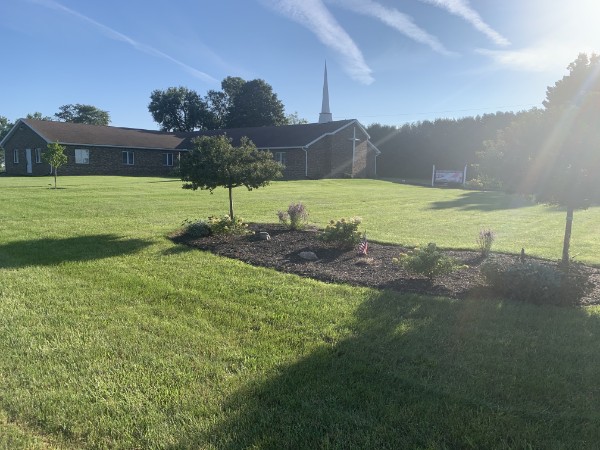 Faith Baptist Church across a green lawn with a landscaped flower bed and white steeple.